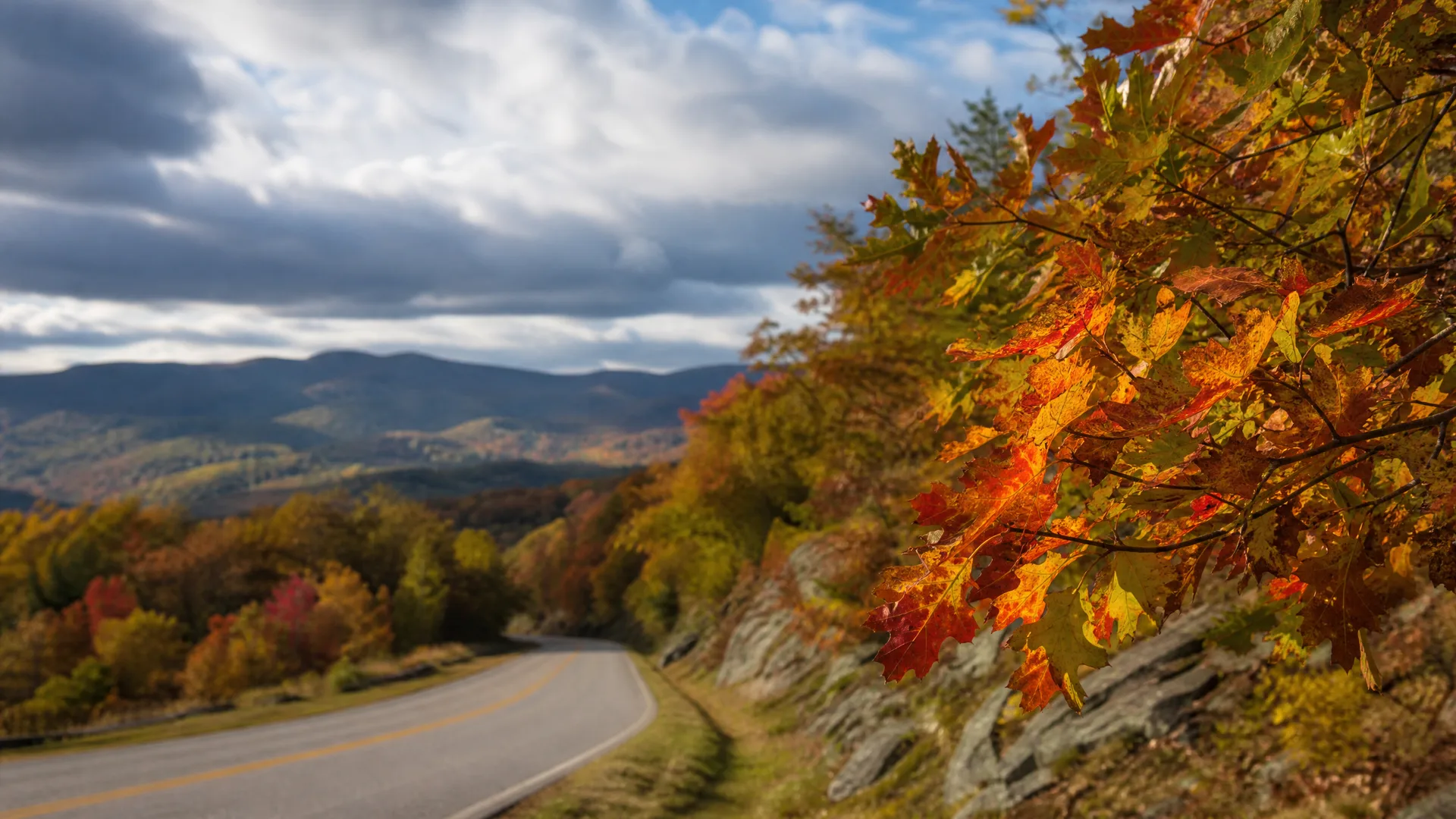 A two-lane road with yellow center lines curves along a ridge past rocky banks and trees in autumn color, with distant forested mountains and a cloudy sky.